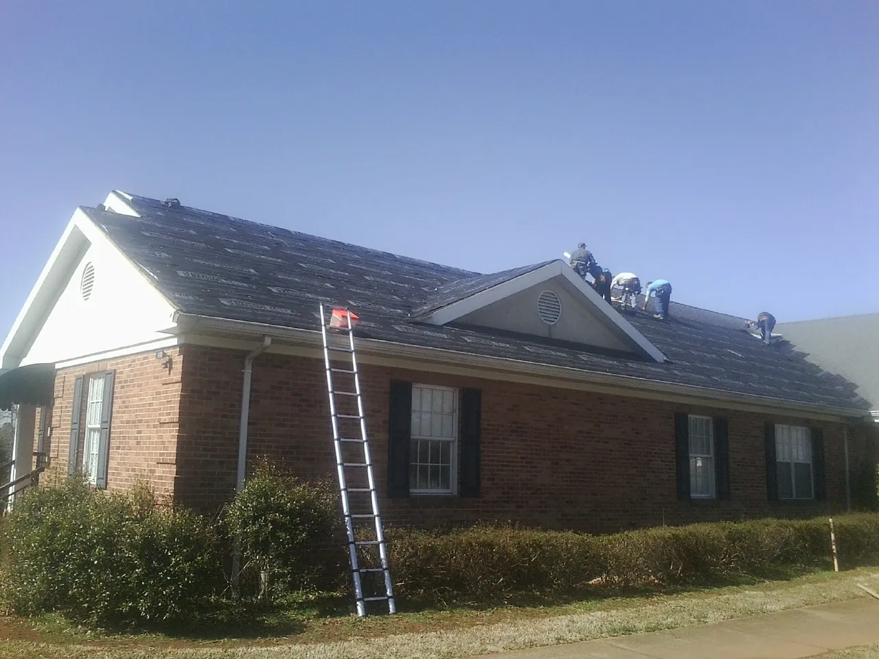 Crew tearing shingles off a brick ranch roof with a ladder set — Metal Roof Repair work by our crew in Dent